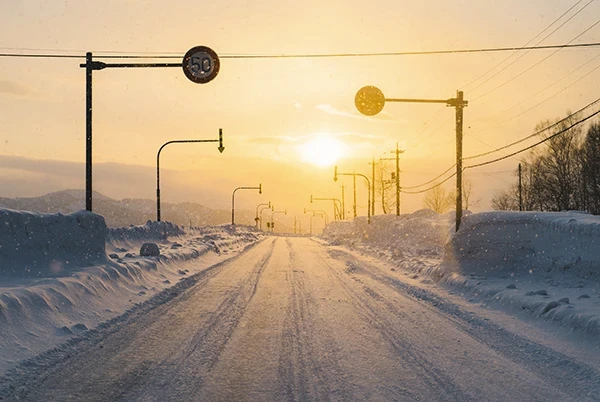 Zimowa droga o zachodzie słońca, otoczona wysokimi zaspami śniegu i znakami drogowymi, oświetlona ciepłym światłem