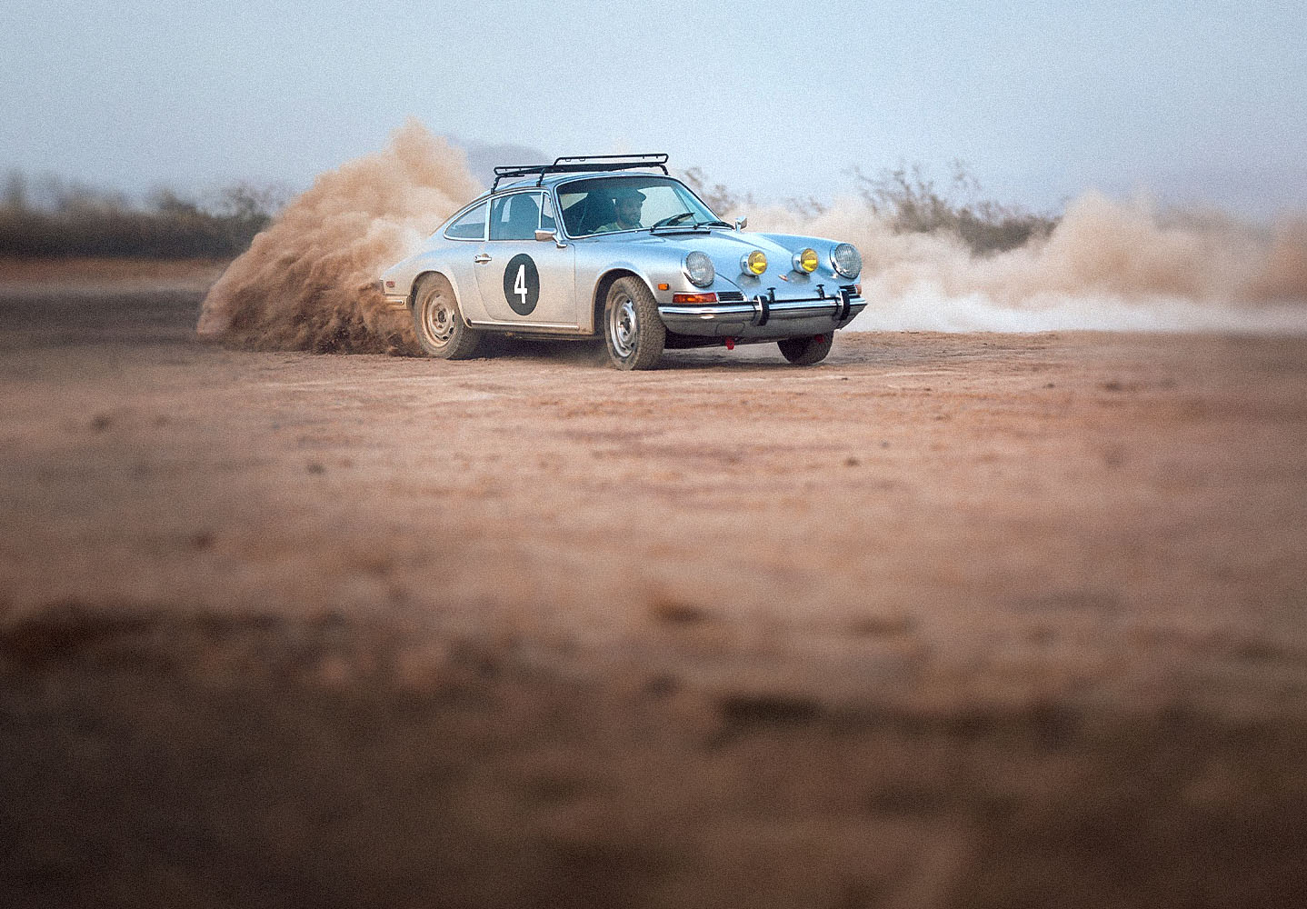 A vintage silver Porsche rally car with the number 4 on its side kicks up a cloud of dust as it drifts on a dirt track in a desert landscape.