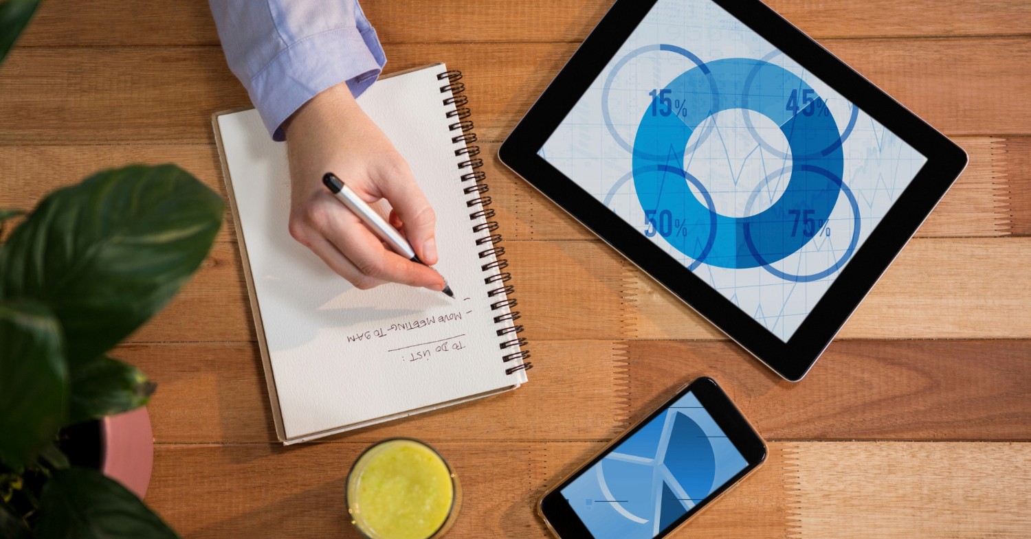  A close-up of a woman's hand writing in a notebook, with a tablet and smartphone placed on a wooden table.