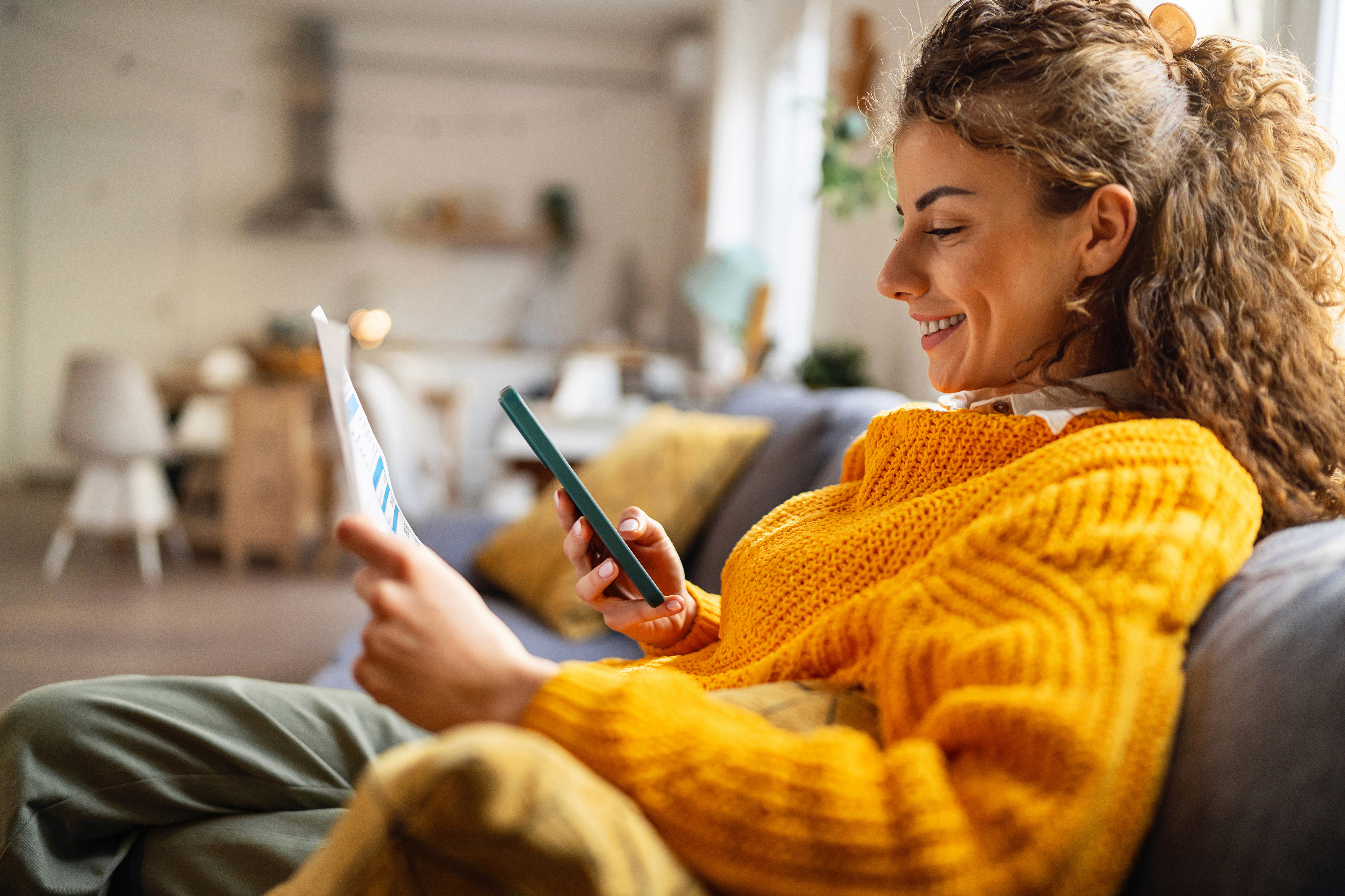 A woman in a cozy yellow sweater sits smiling on a comfortable sofa, holding a smartphone and a tablet in a warmly lit living room.