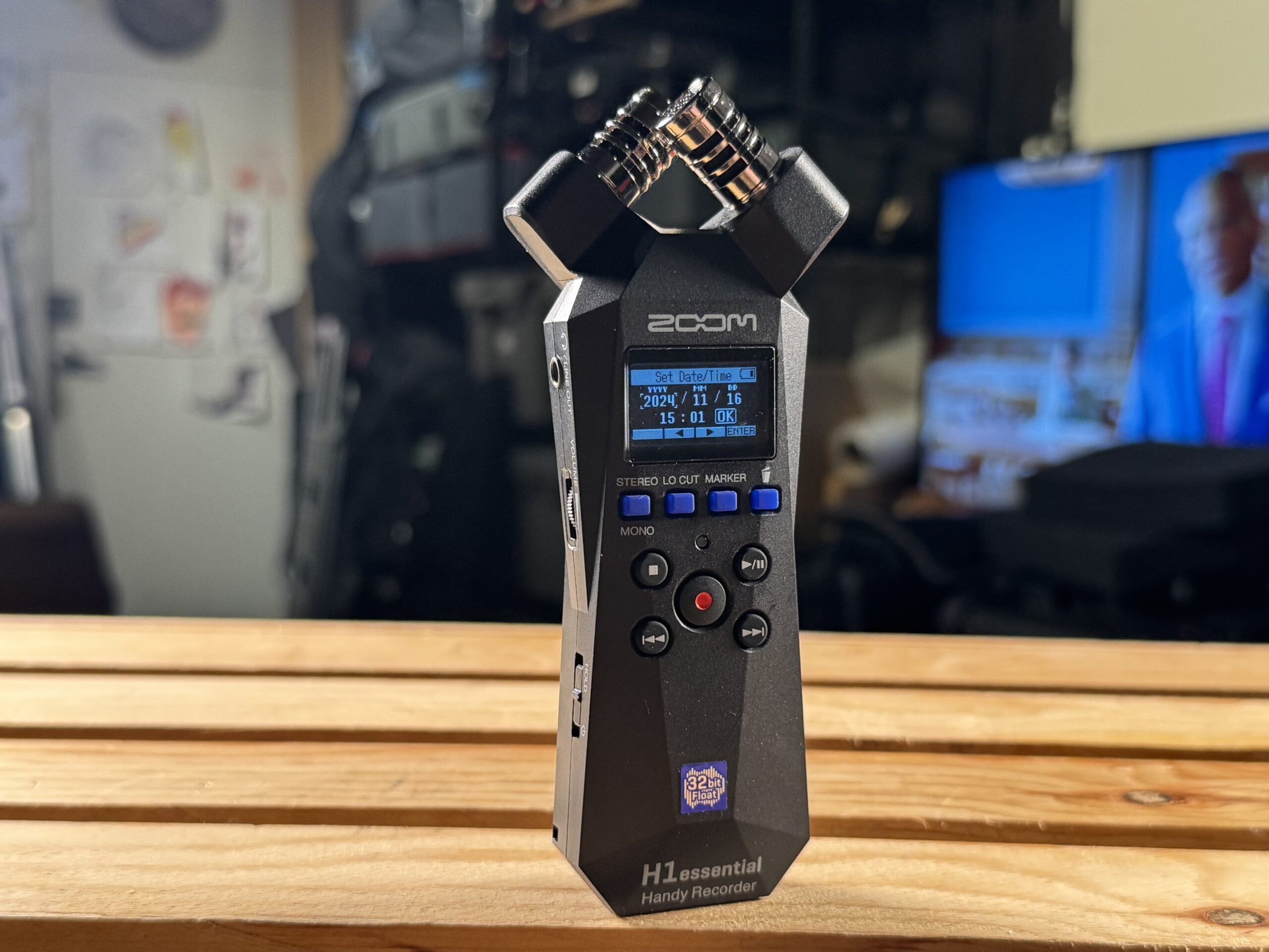 Front view of a Zoom H1 Essential audio recorder standing upright on a wooden table in a studio.