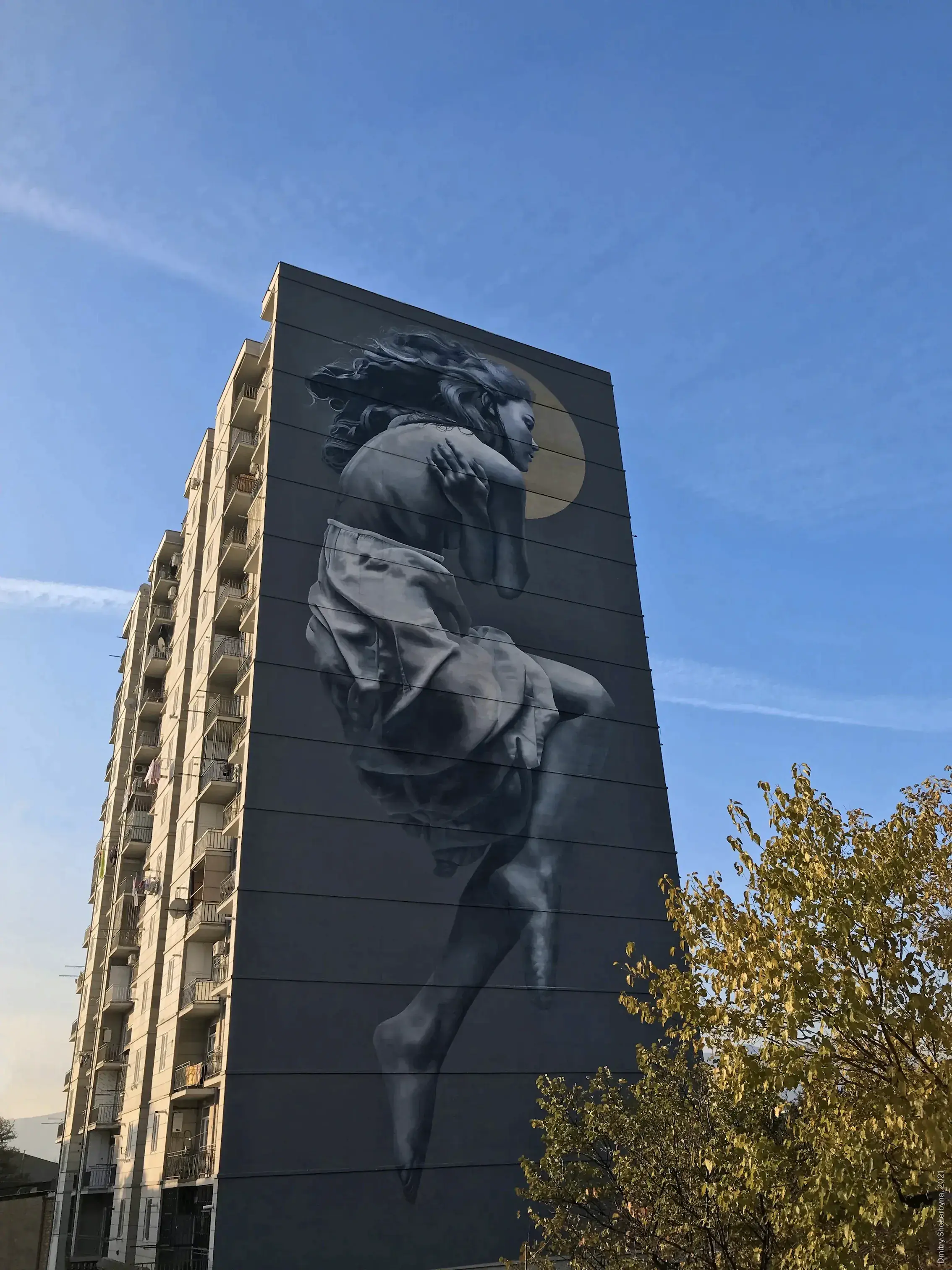A tall mural of a woman on a dark building wall painted in greyscale. She is floating or leaping, with her hair blowing upward. Her hands are pressed together. A golden crescent moon glows behind her shoulders.