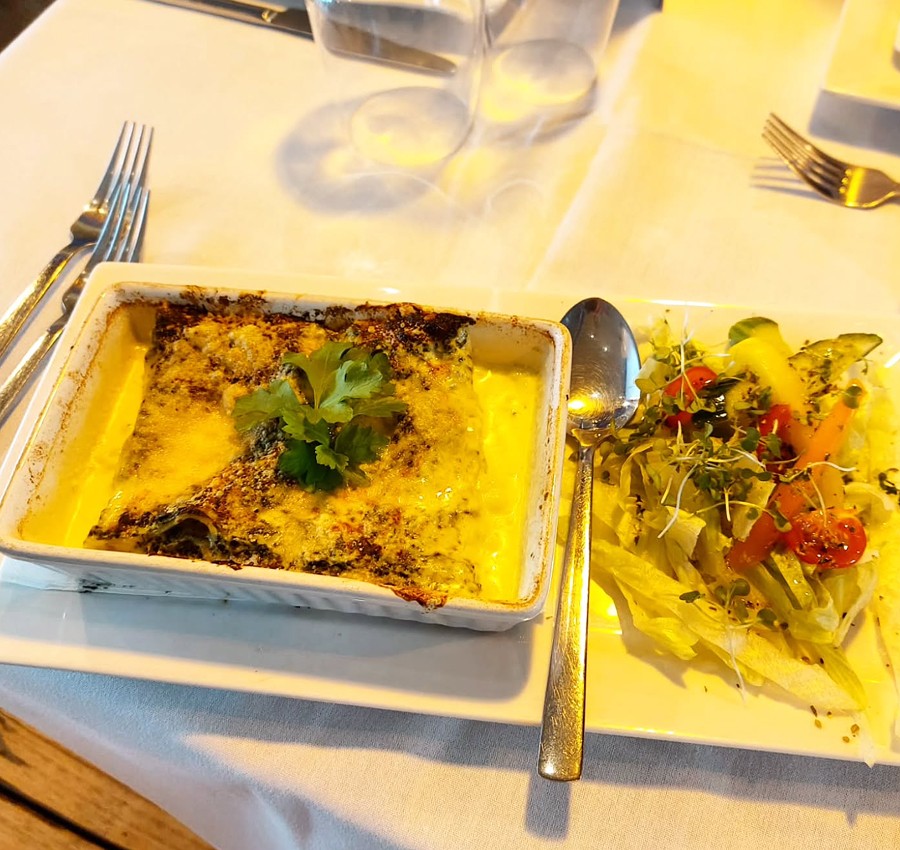 A rectangular dish of baked lasagna topped with melted cheese and a sprig of parsley, served on a white plate alongside a colourful side salad with mixed greens, tomatoes and sprouts, set on a white tablecloth with cutlery arranged nearby.