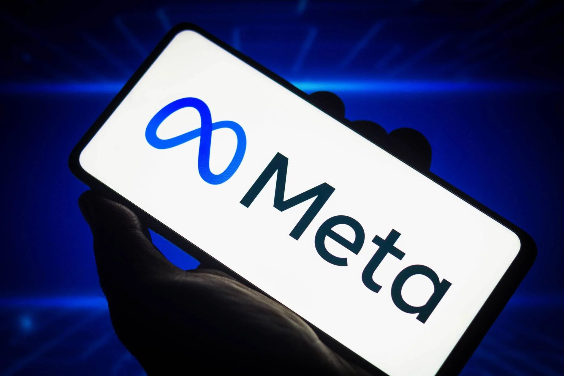 A close-up of a hand holding a smartphone displaying the Meta logo on its screen, set against a solid blue background.