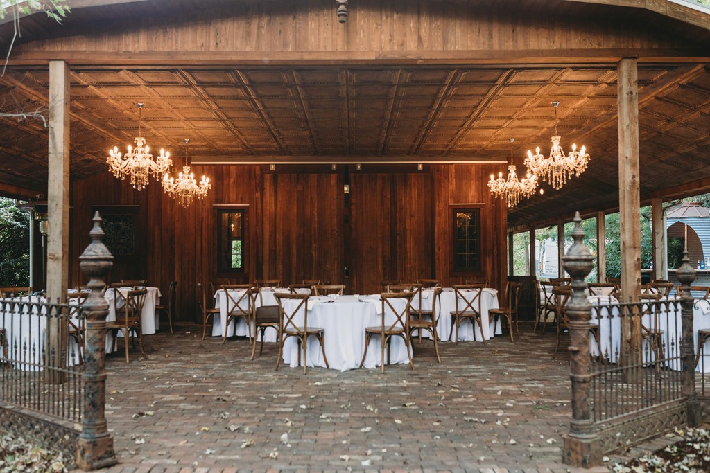 Covered rustic wedding reception space with crystal chandeliers, round white-clothed tables, crossback chairs, and a brick floor patio