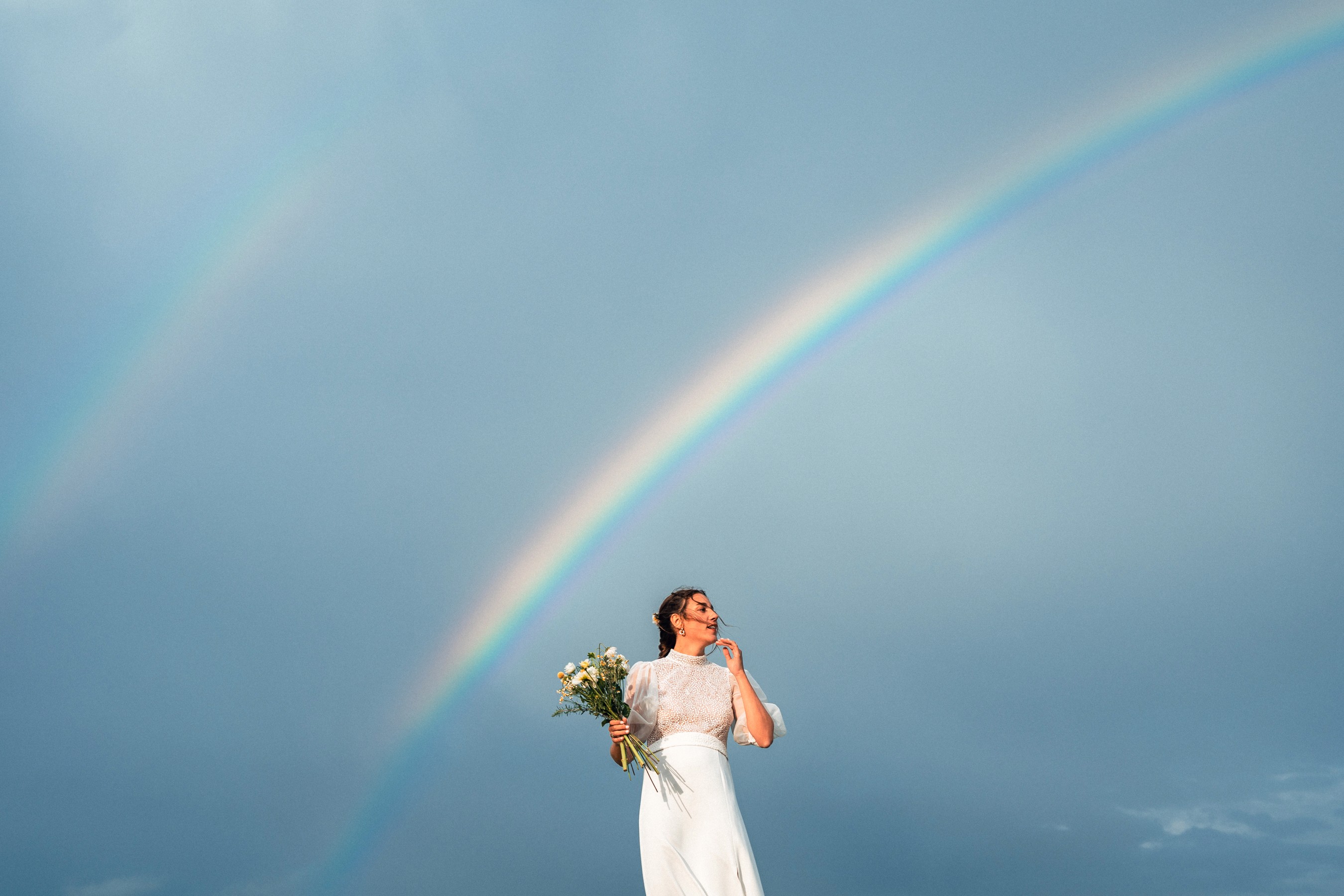 Photographe de mariage au Pays basque, bouquet lancé dans le ciel sous un arc-en-ciel, symbolisant une vision humaine, intense et poétique du mariage.