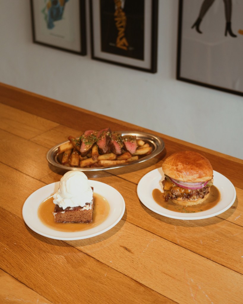 A table displaying a dessert of cake topped with ice cream and sauce, alongside a burger with toppings and a platter of sliced meat with fries in the background.