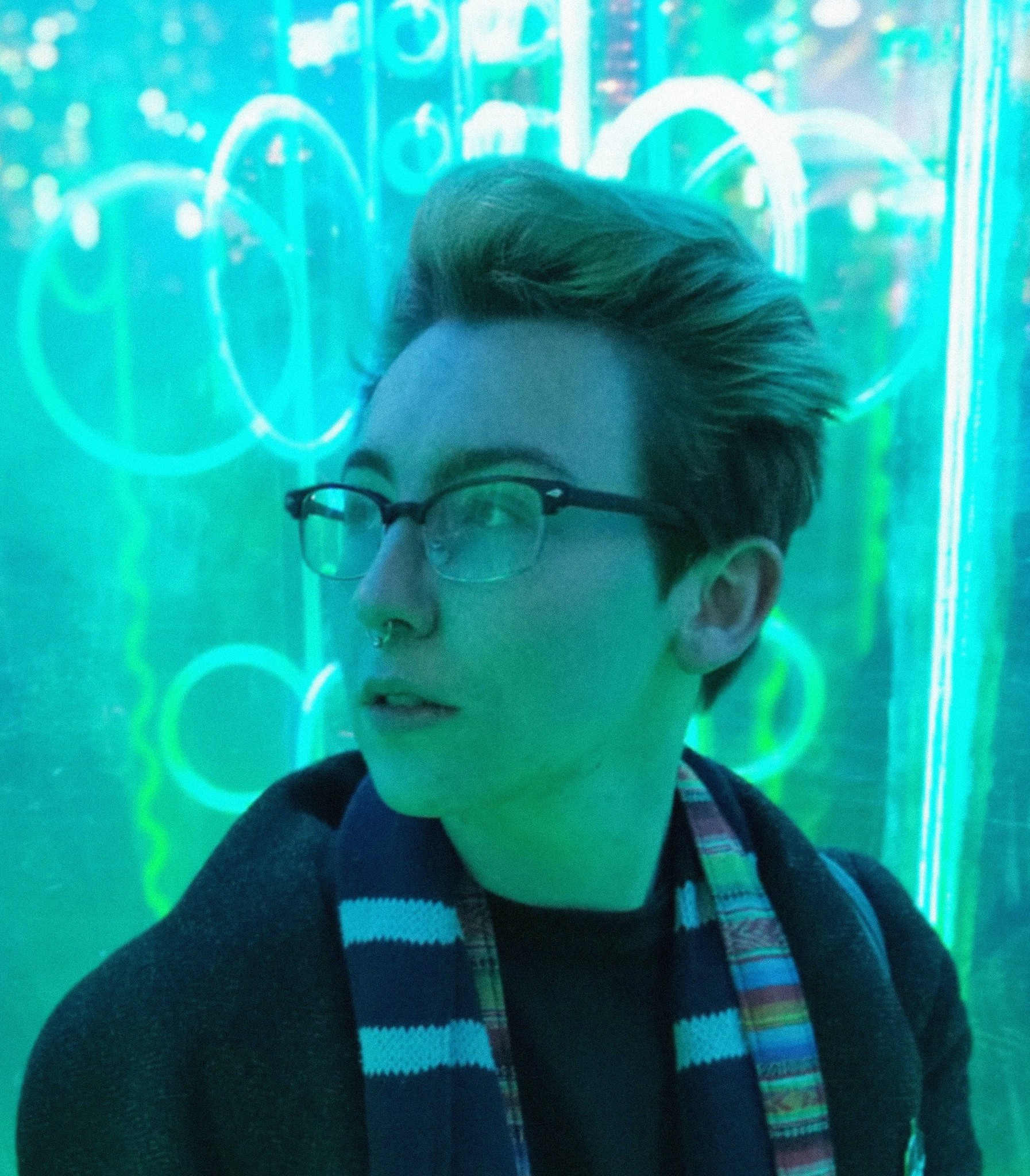 A photo of Ruben, a trans masculine individual with short hair and glasses. He is in 3/4 profile, looking off to the left. Behind him are a number of neon lights, which reflect and turn the whole image teal.