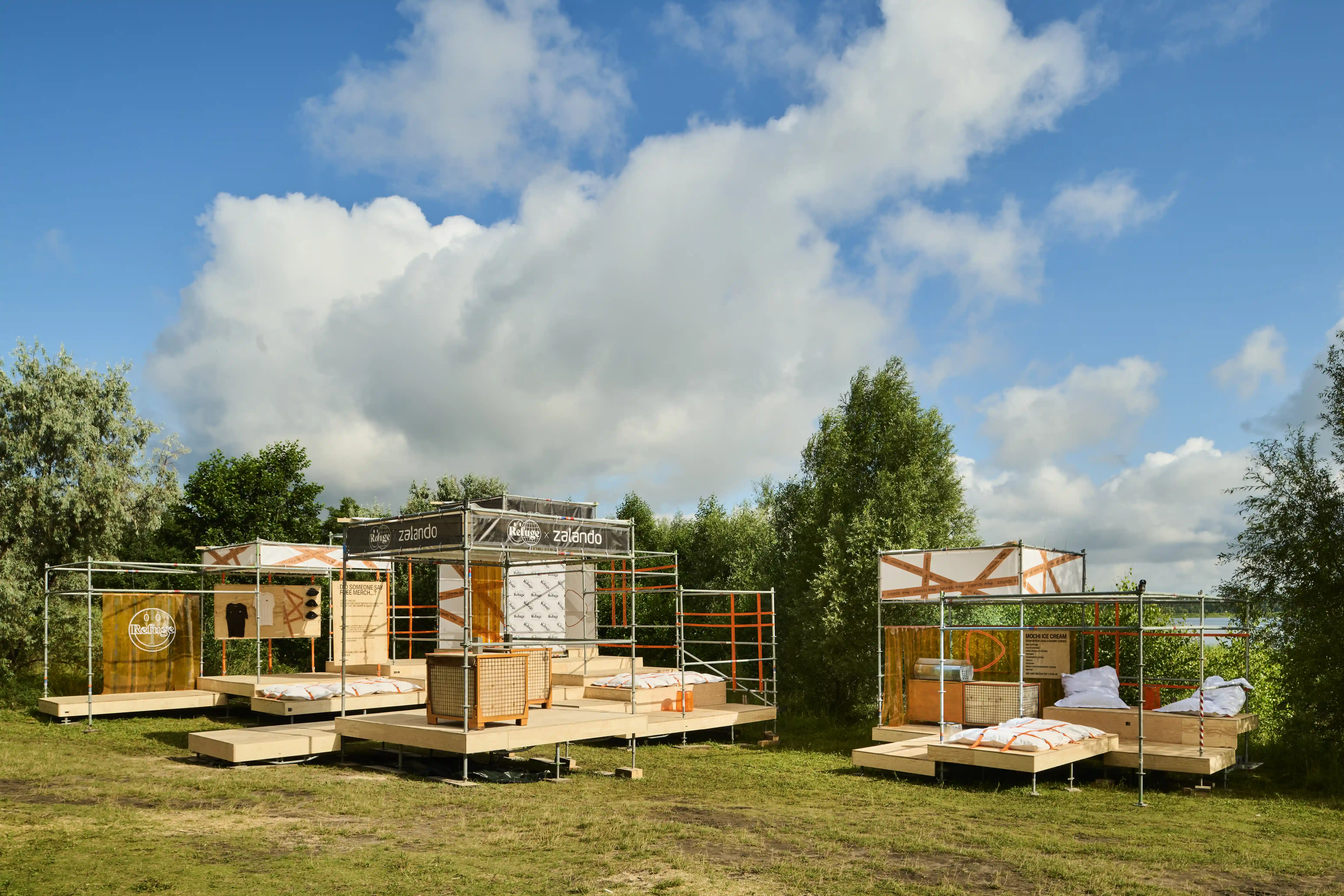Festival installation of scaffold pavilions with wooden decks and beds in a green meadow.