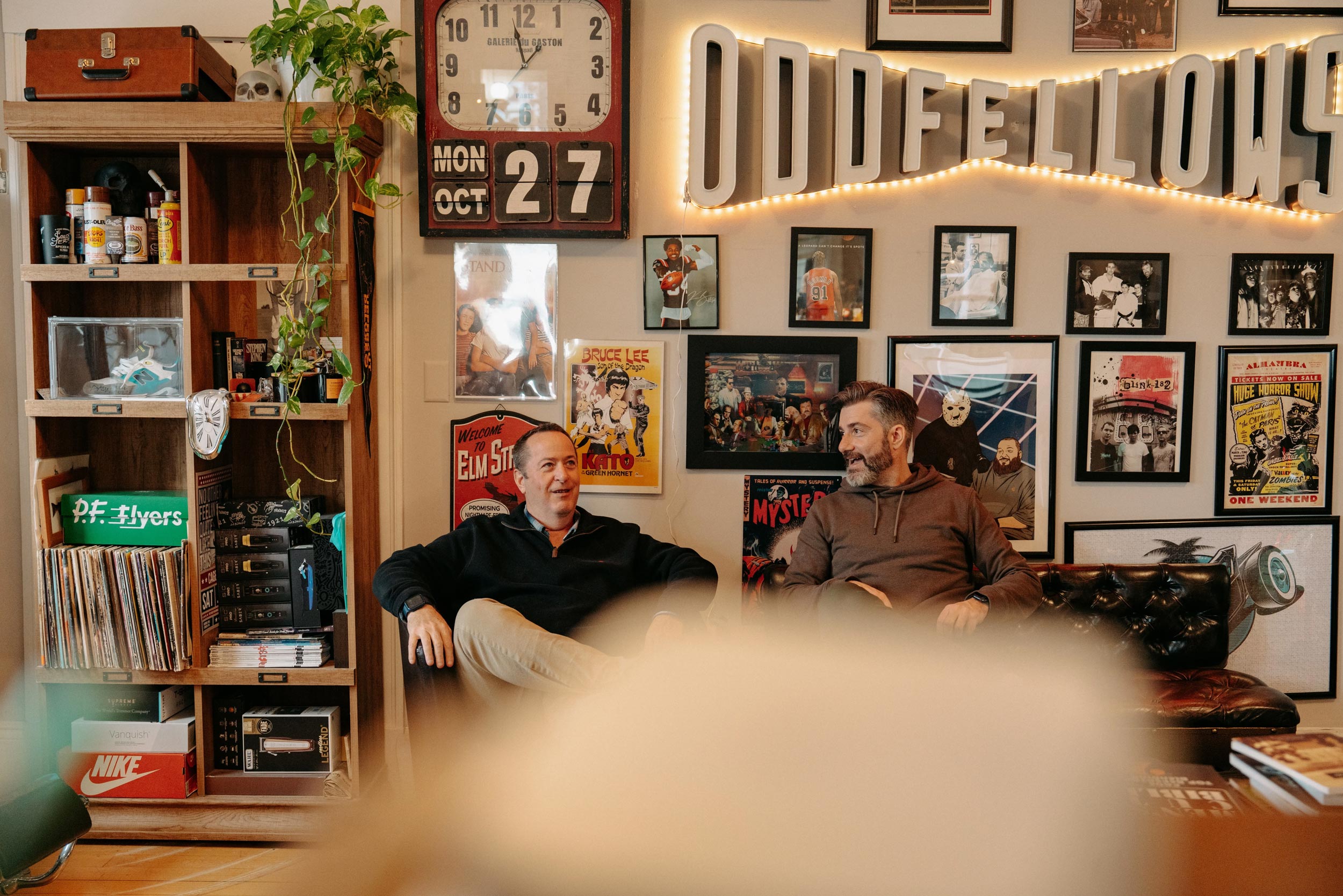 An interior view of a retro barbershop, "ODDFELLOWS," featuring a large, lighted sign with its name. Two men are seated on a couch in the waiting area, with a wall behind them decorated with vintage movie posters, including those for Bruce Lee, A Nightmare on Elm Street, and Stand By Me. A shelf to the left holds a collection of records and a boombox.