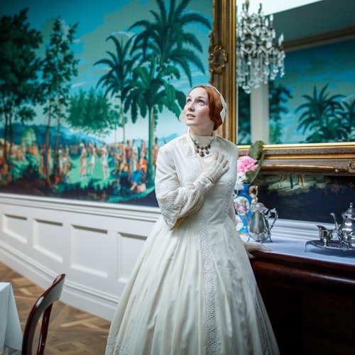 一个穿着白色复古礼服的人站在室内，身旁是一张摆放着银色茶具的桌子，上方悬挂着一幅大型棕榈树画作。