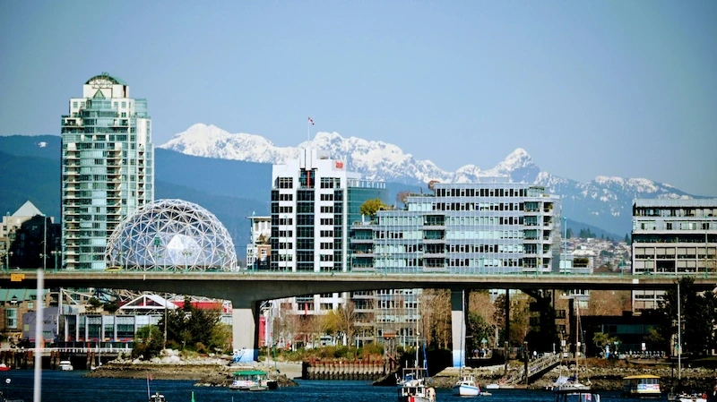 False Creek waterfront near Mount Pleasant with Science World dome, modern buildings, boats, and snowy mountains.