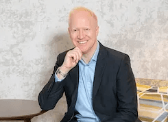 Professional headshot of Simon Zutshi wearing a dark suit jacket and light blue shirt, smiling at camera