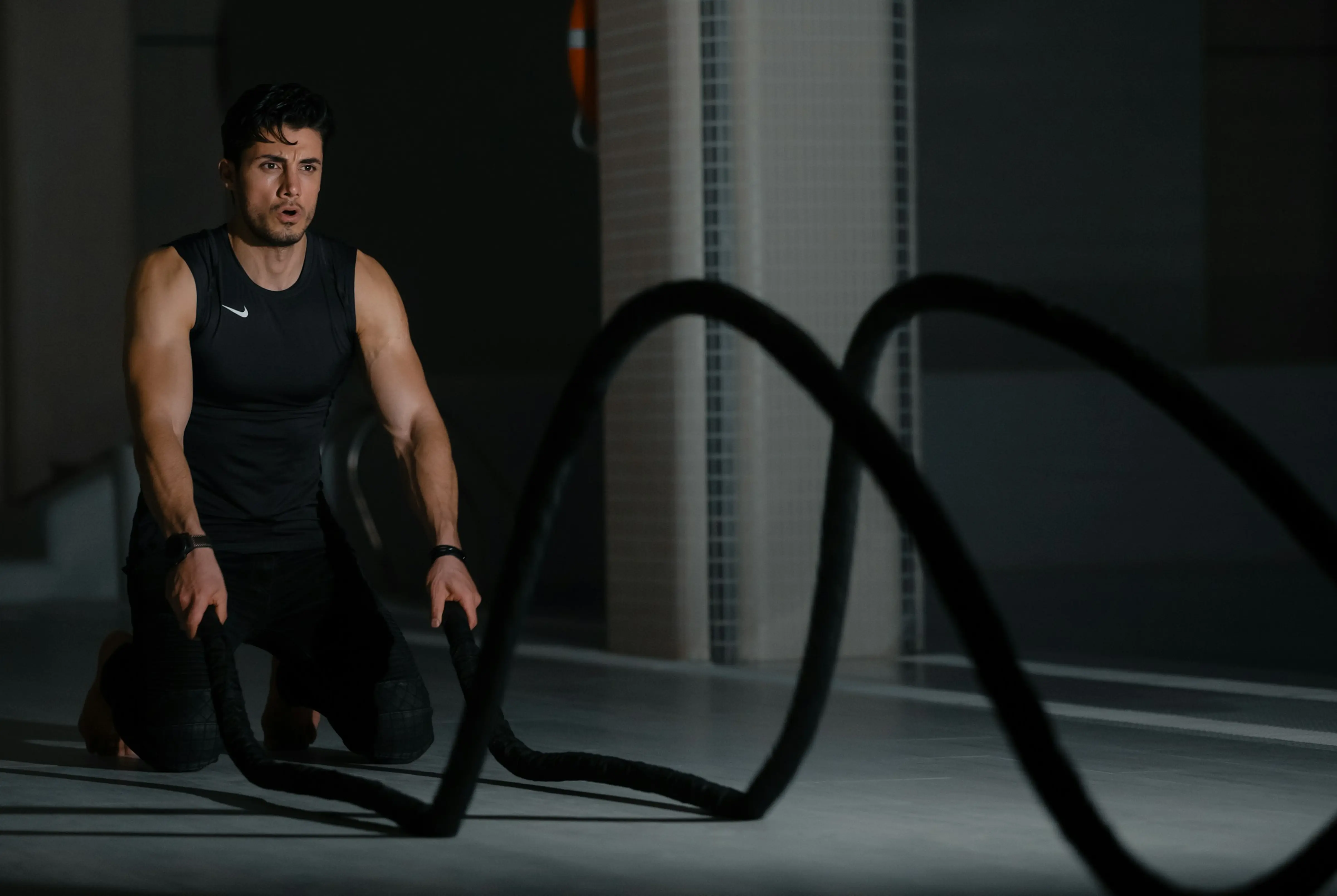 Fit man kneeling on the gym floor performing battle rope exercises in a dark training space.