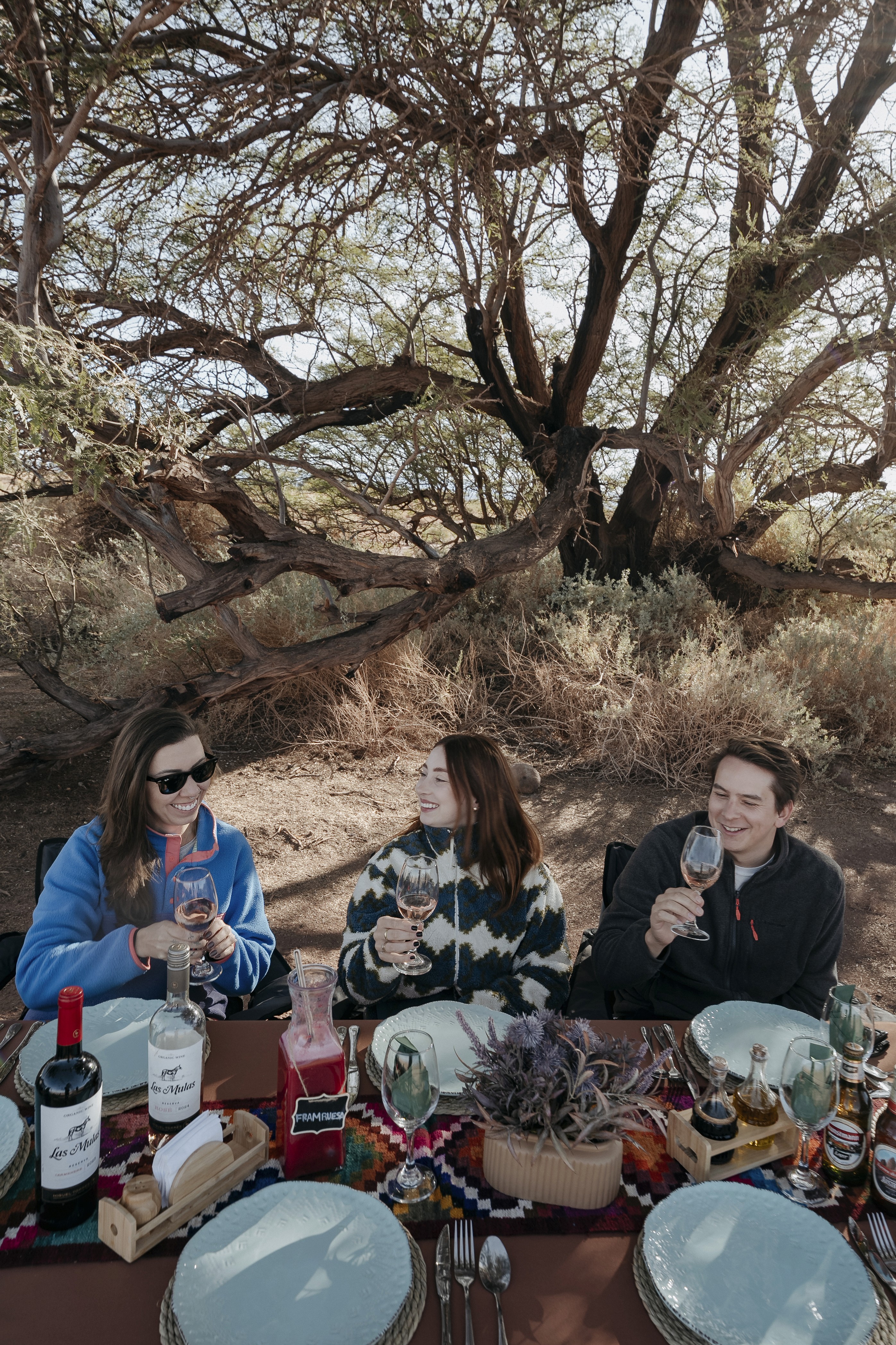 A young person with curly hair smiles at the camera while sitting in front of a laptop against a dark background.