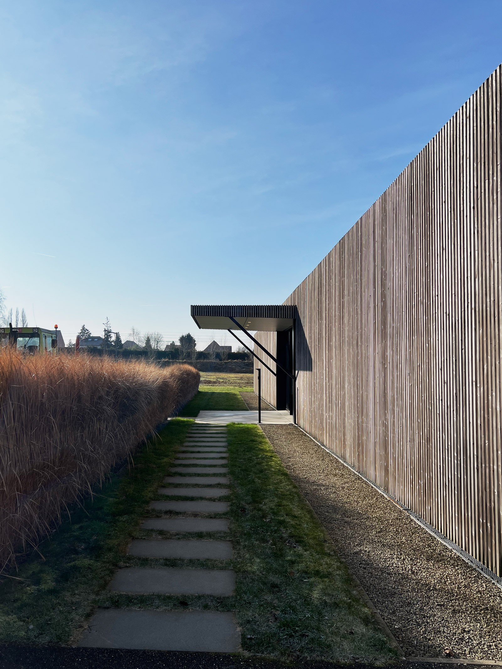 Een modern architecturaal detail van een gebouw met een verticale houten gevelbekleding. Een strak zwart afdak markeert de ingang, met een pad van betonnen tegels dat door het gras langs de gevel loopt.