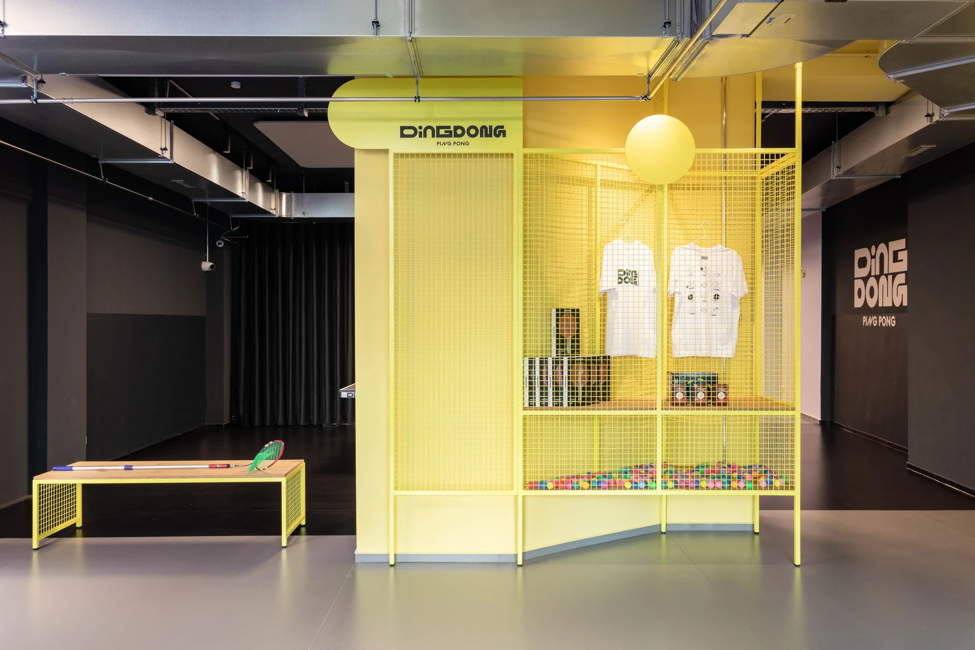 DingDong merch zone in bright yellow with mesh display walls, branded t-shirts on hangers and a colourful ball-pit beneath the shelving.