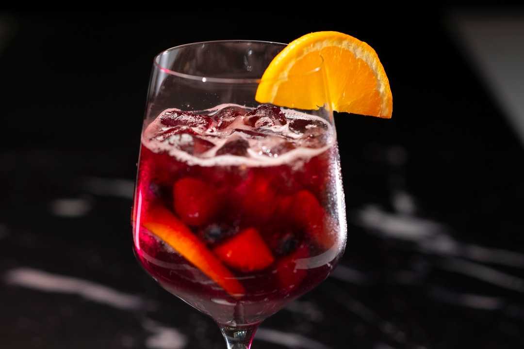 A red wine sangria cocktail filled with ice and fresh sliced citrus fruits, garnished with a bright orange wheel in a stemless wine glass.