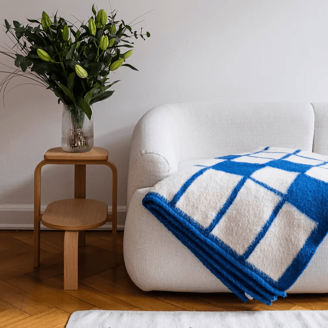 A cozy living room corner features a modern white sofa draped with a blue and white checkered blanket, accompanied by a wooden side table holding a vase with green leafy plants, set against a warm hardwood floor.