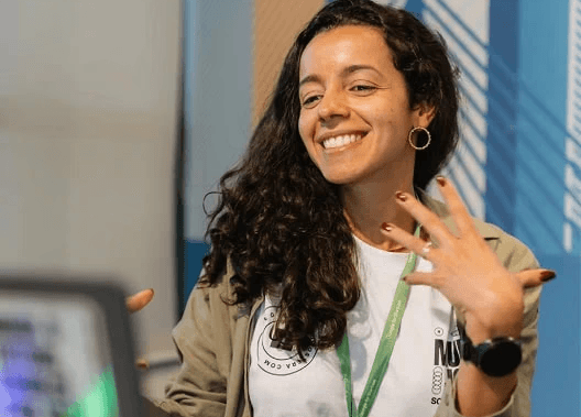 Uma mulher sentada em uma mesa, concentrada em seu laptop, com plantas ao fundo e um interior casual e moderno.