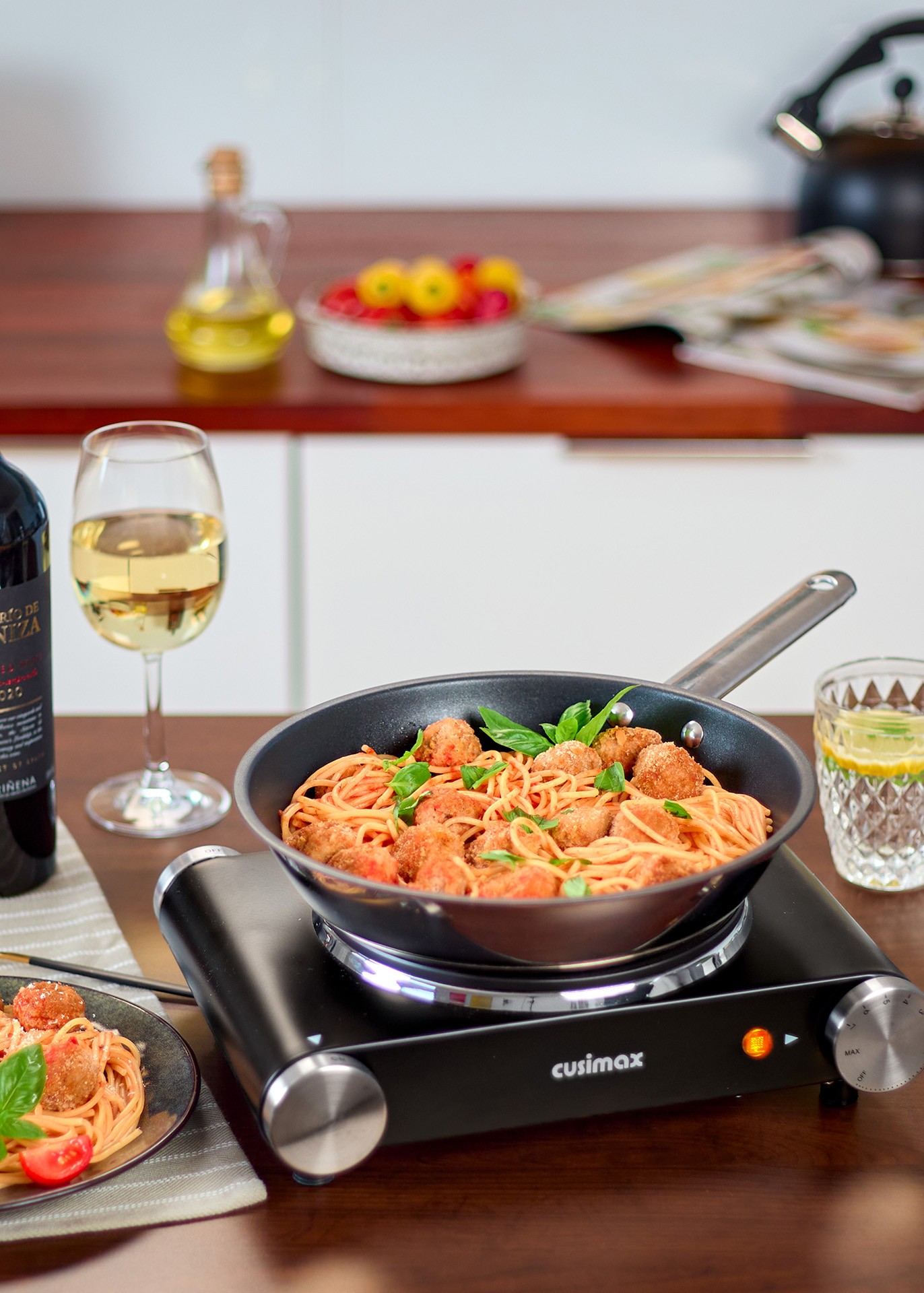 A frying pan filled with spaghetti and meatballs garnished with fresh basil sits on an induction cooktop, surrounded by a glass of white wine, a glass of water with lemon, and a plate of pasta on a wooden kitchen counter.