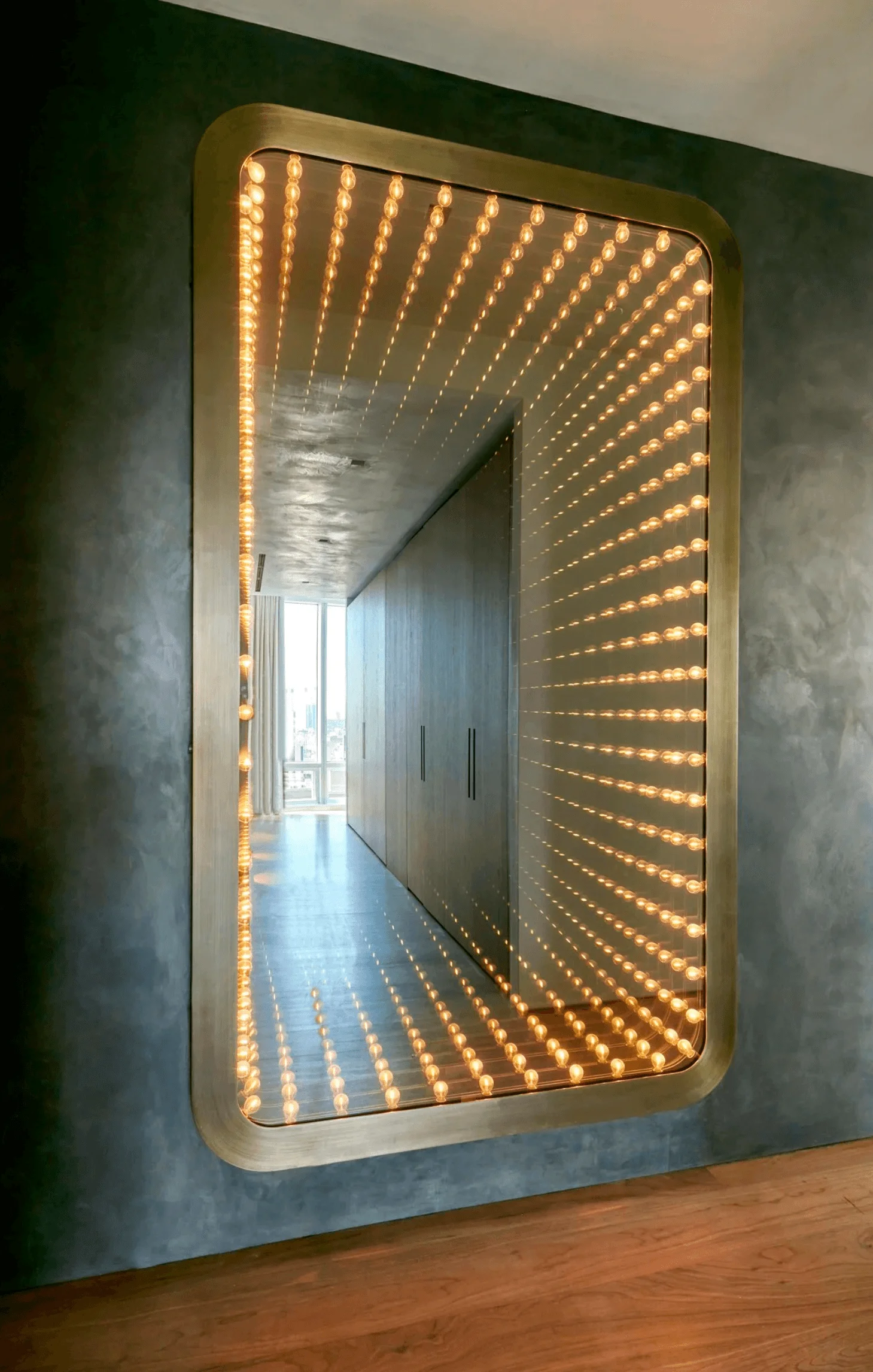 A brass-framed light-bulb infinity mirror hangs on a textured gray wall opposite a wood-paneled hallway