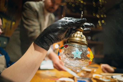 A person crafting a Turkish mosaic lamp, carefully applying multicolored beads to the ornament. The background shows a workshop setting with tools hanging on the wall.