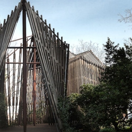 Drewniane konstrukcje z pionowymi belkami ułożonymi w trójkątny wzór, otoczone drzewami, pod bezchmurnym niebem.