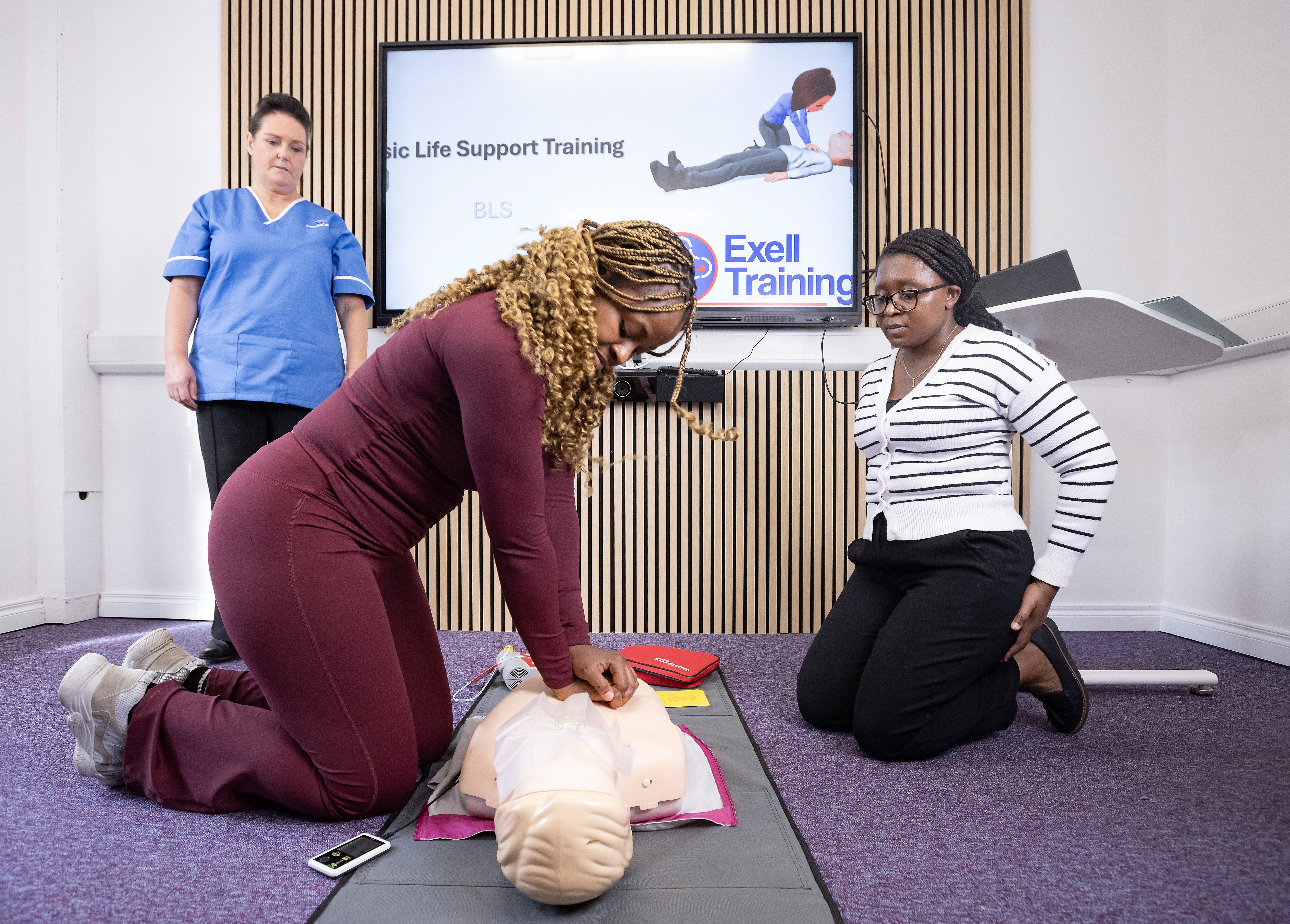 A person performing CPR on a CPR Dummy. Two other people supervising and watching