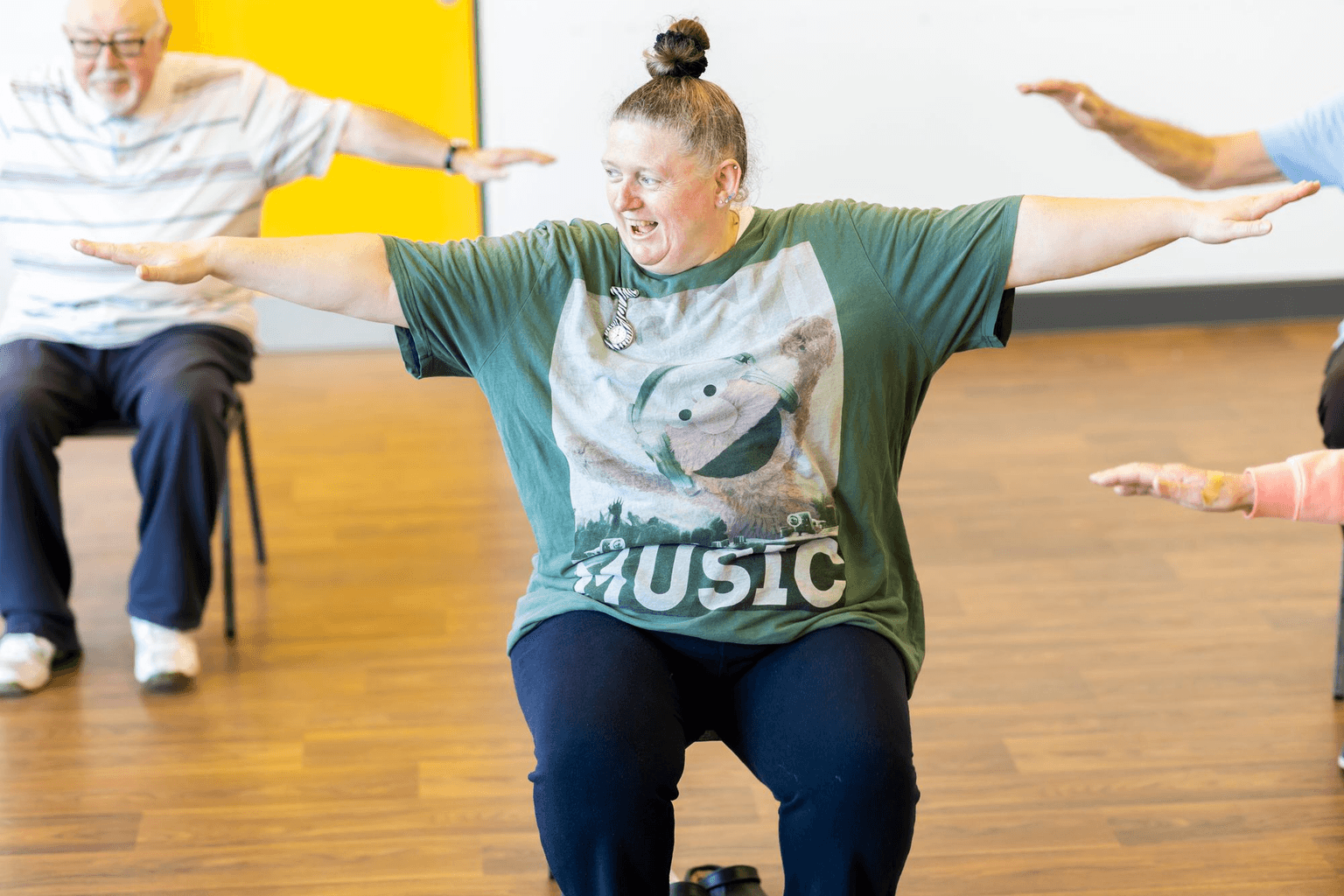 A seated exercise class mid movement, the instructor leading with both arms out.