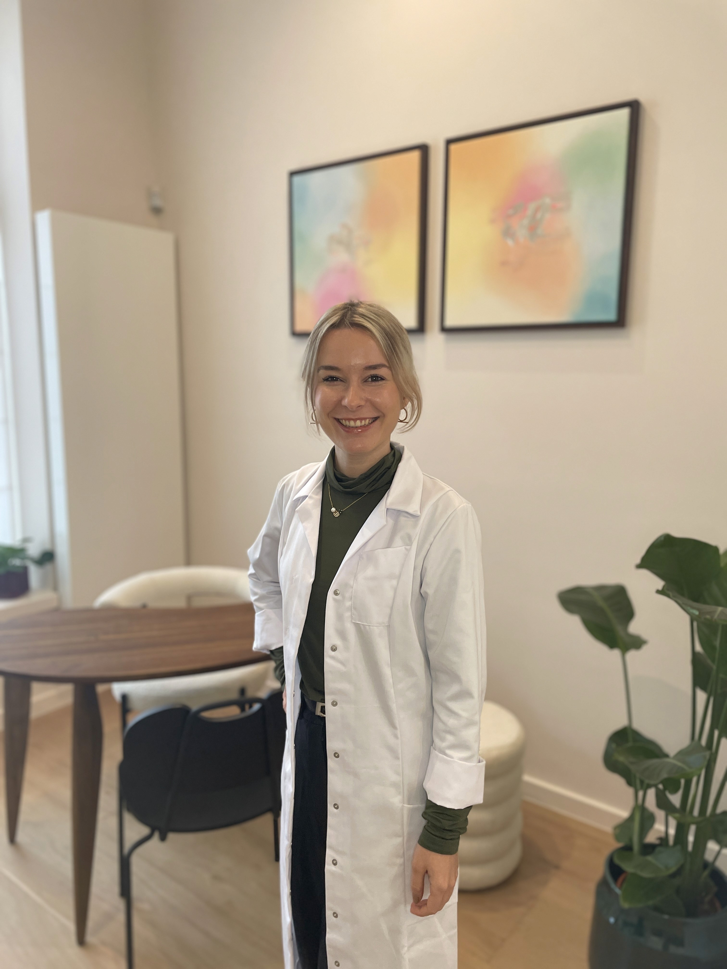 Dr. Louise Cuveele standing in her practice, with two colorful paintings in the background, a desk with chairs for consultations, and a visible plant.