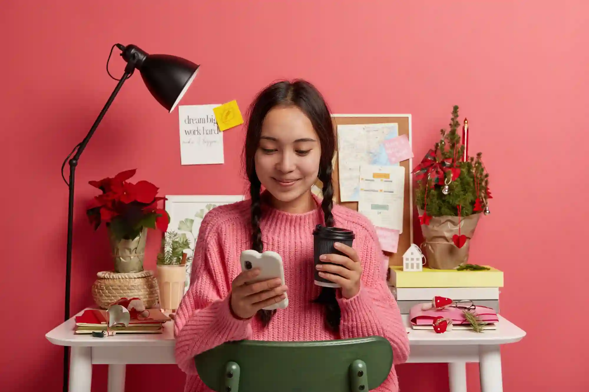 A young woman smiling and checking her phone while holding a coffee cup at a festive desk setup.