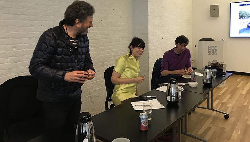 A photo of three persons standing near a desk and giving a lecture.