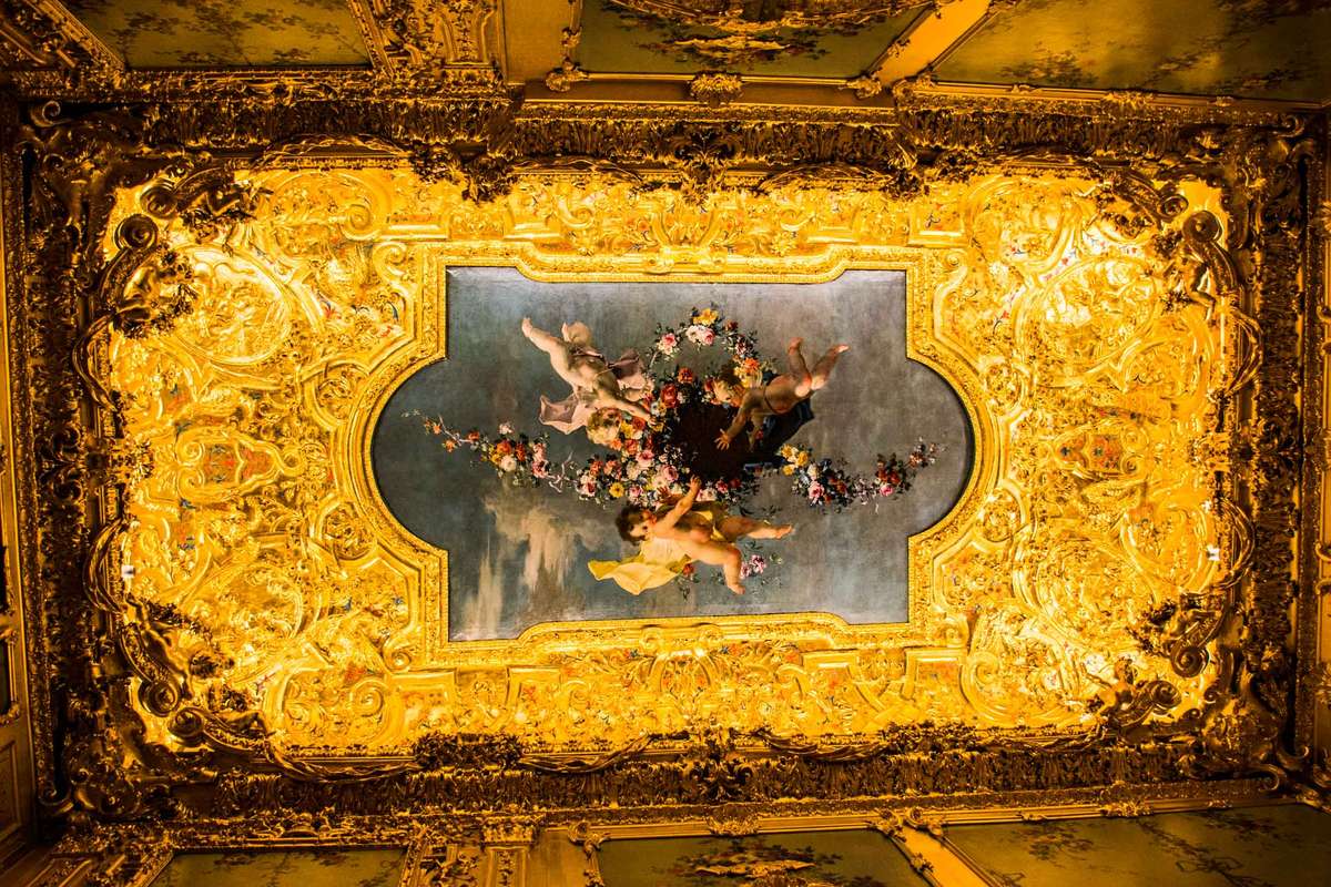 A gold-leafed ornate ceiling features a central rectangular painting of figures in classical attire against a blue background.