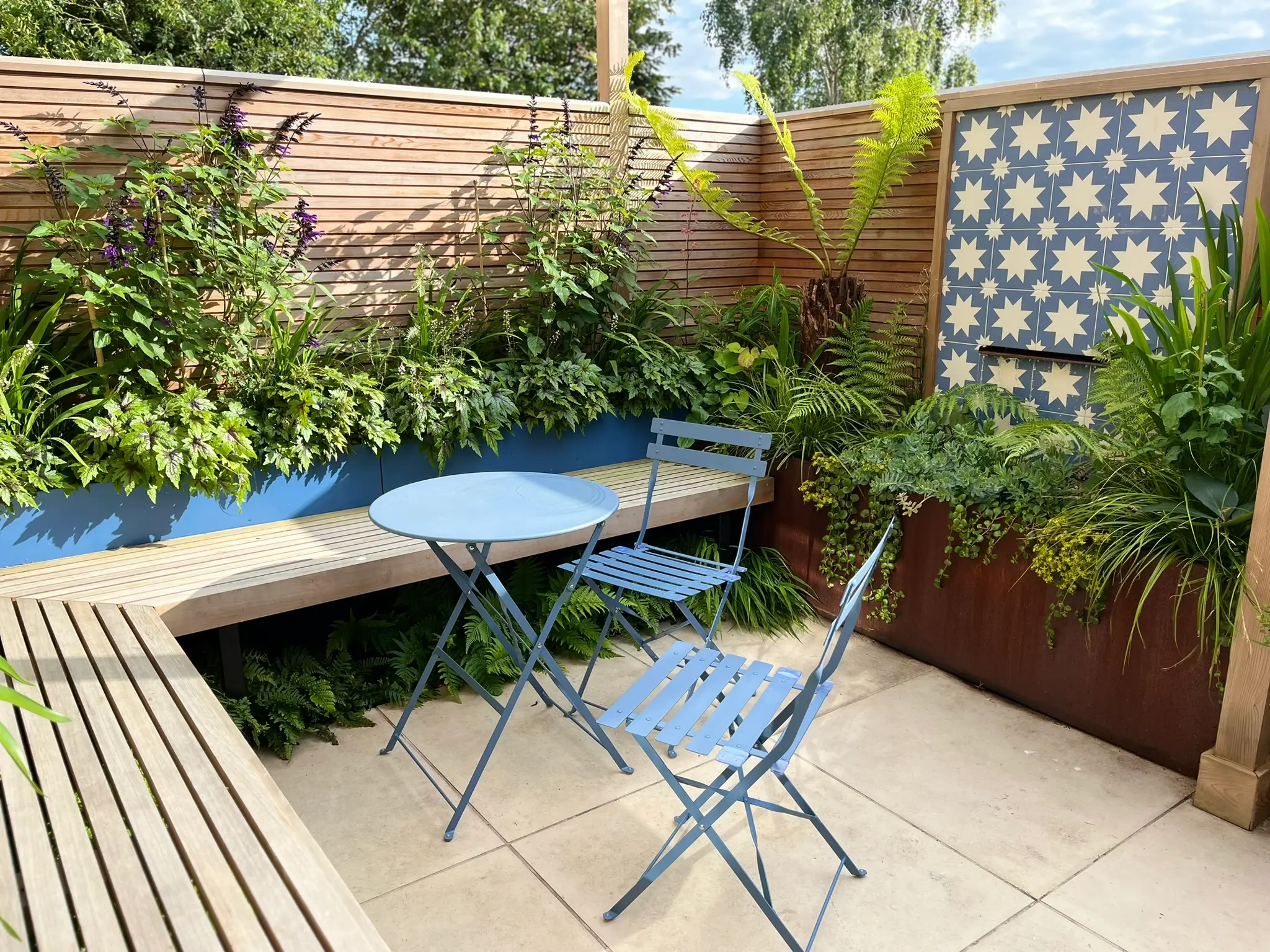 A cozy outdoor patio featuring a wooden table with blue chairs, surrounded by lush greenery and decorative wood panels.