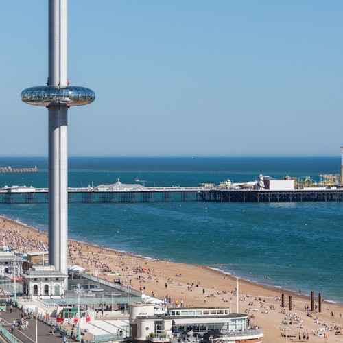Beach with scattered people, a pier extending into the sea, and a tall observation tower with a glass observation deck.
