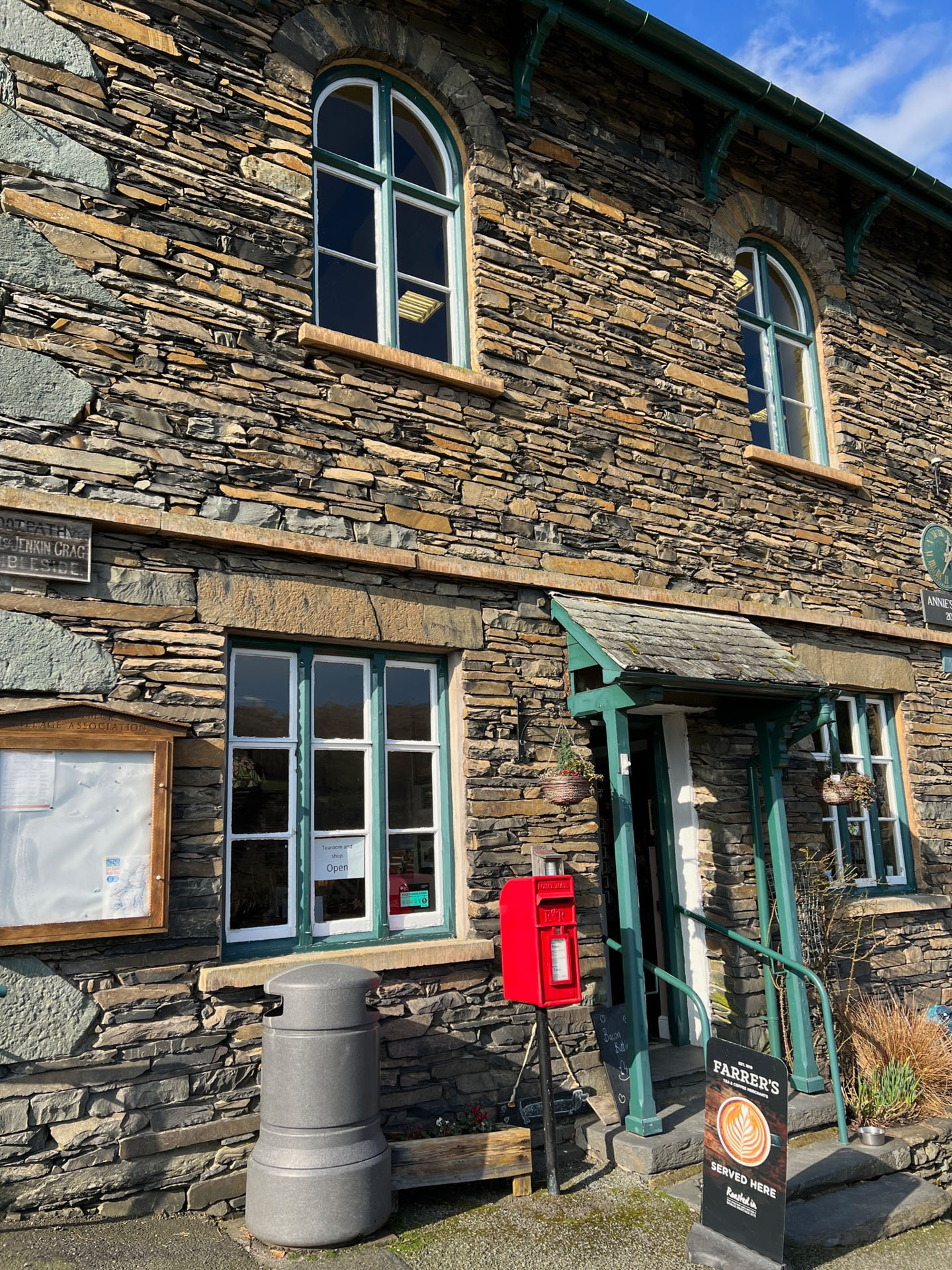 An old stone building with single-pane windows. A bright red post box is situated outside by the front door, along with a Farrer's Coffee sign. The woodwork of the building is a teal-green colour.