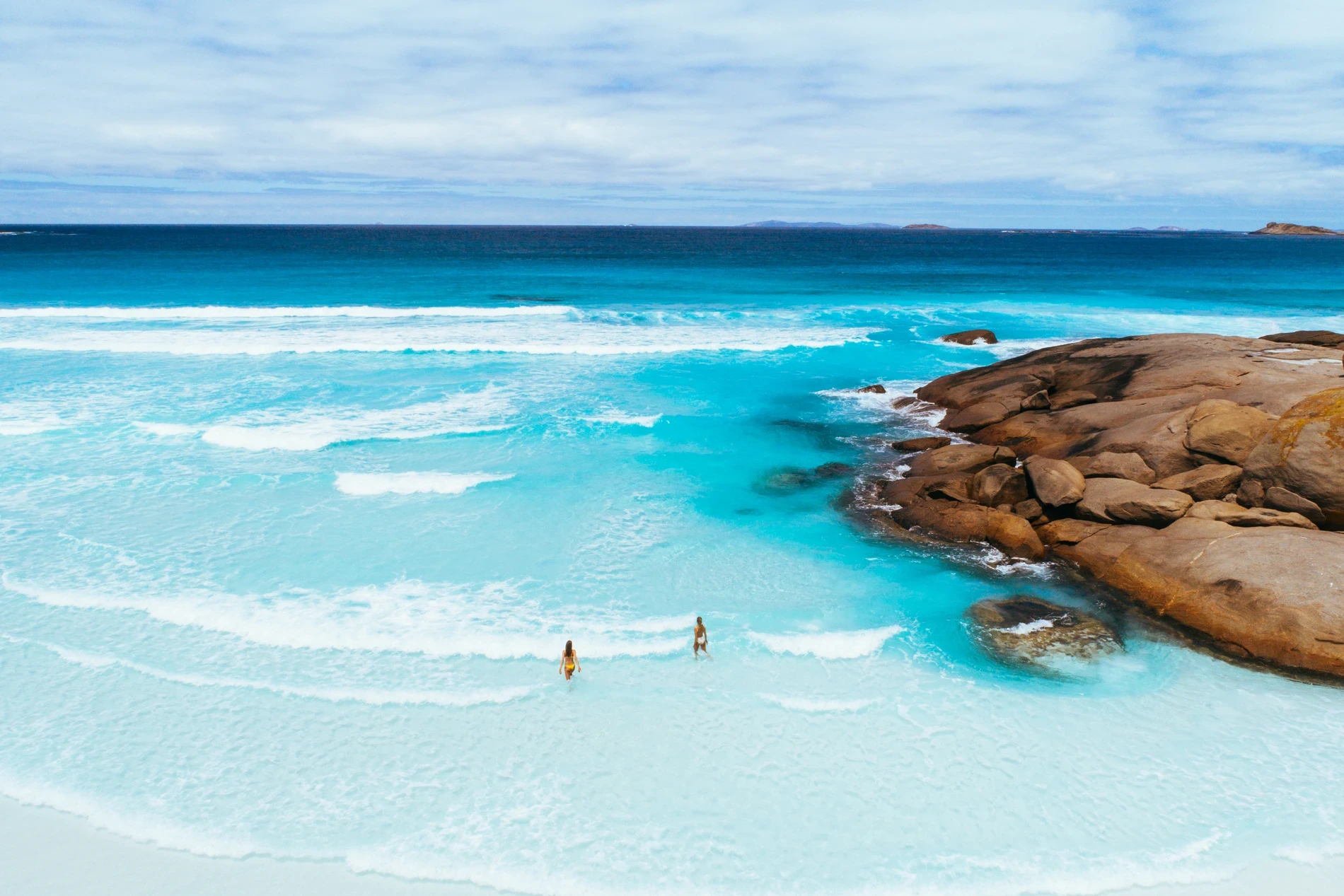 Two people wading in turquoise shallows beside granite rocks and breaking surf