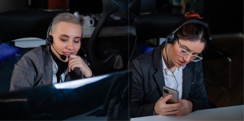 Two customer service representatives in an office setting wear headsets; one is engaged with a computer monitor while the other checks a smartphone, both showcasing professional business attire.