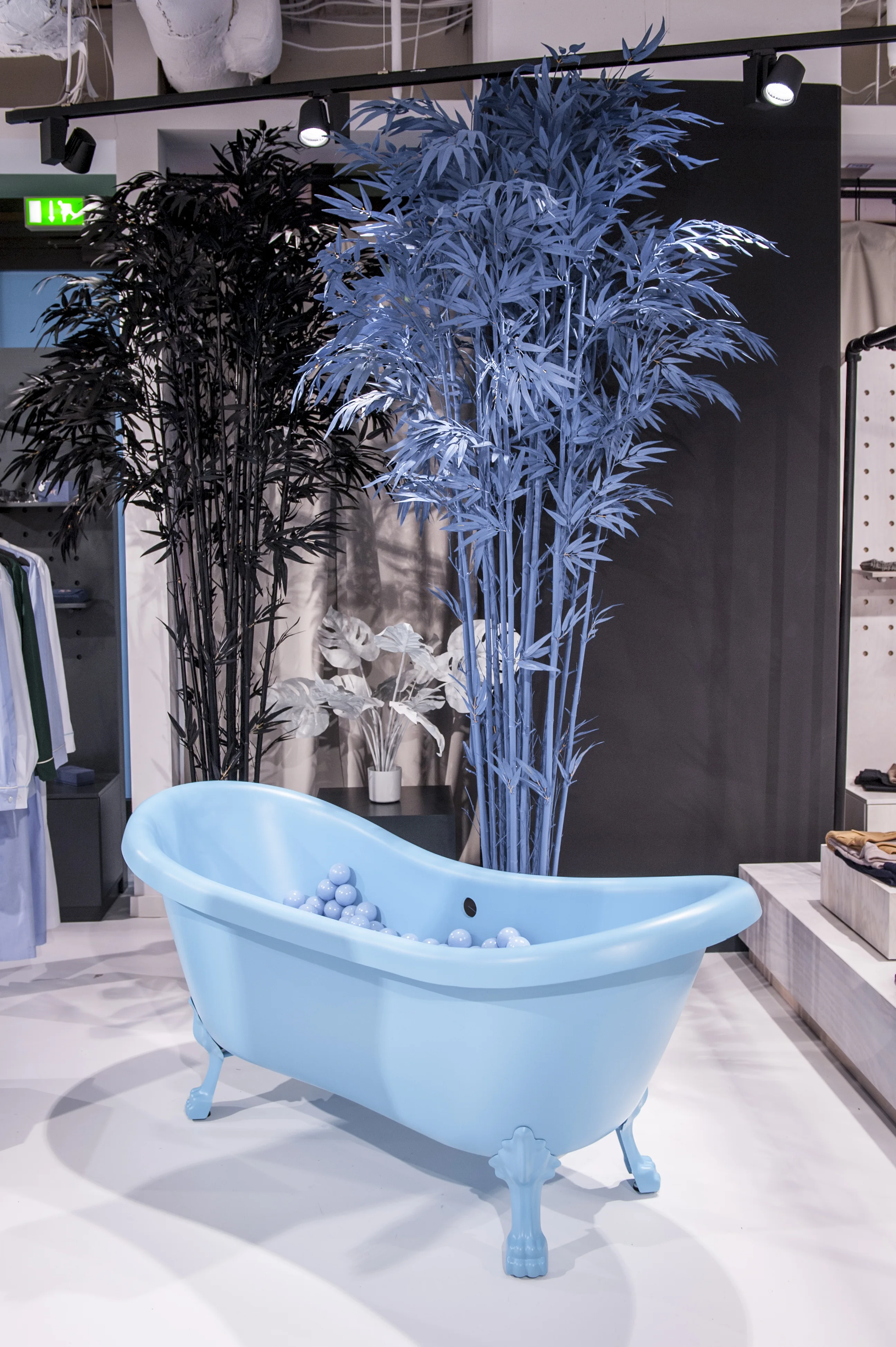 Light blue claw-foot bathtub filled with blue balls at FREIRAUM SHOP, set against blue and black painted plants in front of a dark partition.