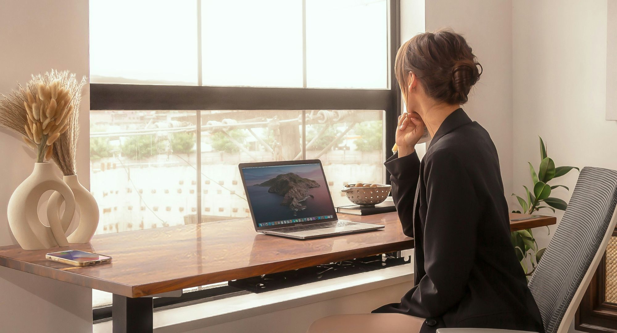 mulher sentada à mesa de madeira com um notebook, a mesa está a frente de uma janela de vidro, a mulher está com  o cotovelo do braço direito na mesa com o queixo apoiado na mão, enquanto olha para fora da janela