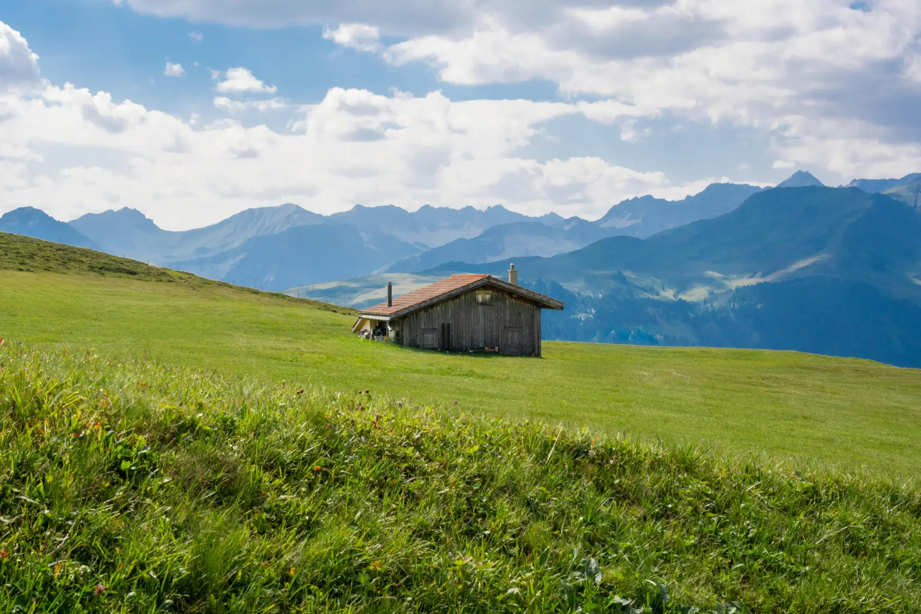 Kleine Holzhütte auf grasbewachsenem Hügel vor fernen Bergen unter bewölktem Himmel.