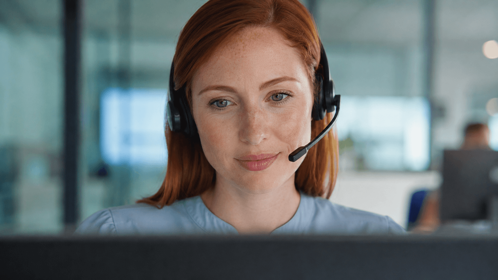 A young female patient-support advisor with auburn hair wears a modern headset while working at her monitor.