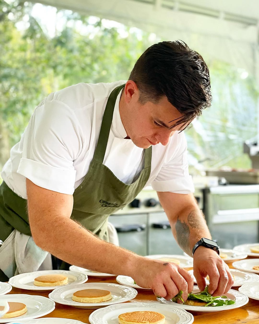 Chef plating food with focused attention. Chef wearing a white shirt and green apron, working on individual plates.