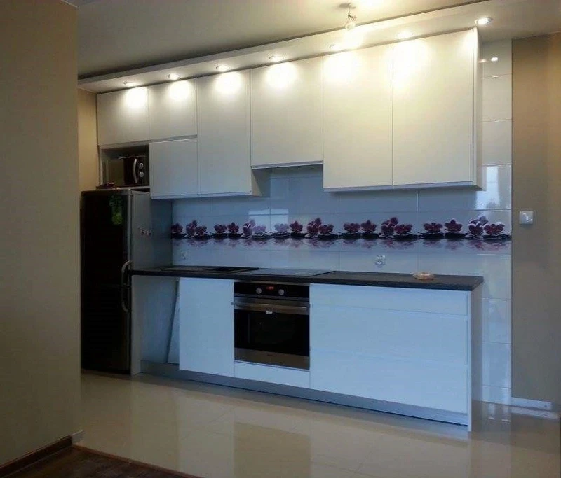 Biała zabudowa kuchenna w wysokim połysku z oświetleniem LED pod szafkami i ozdobnym panelem szklanym z motywem kwiatowym.