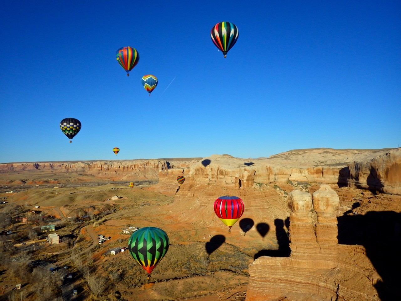 Eight hot air balloons in flight at a balloon festival over red rock cliffs and a small desert town in southern Utah, their shadows falling across the sandstone below.