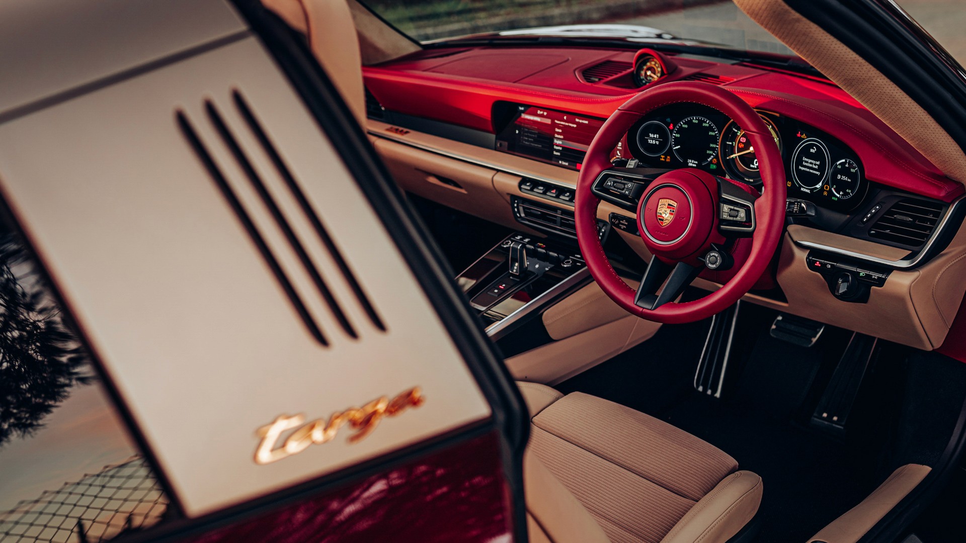 Interior of a Porsche 911 Targa 4S Heritage Design Edition, showing the red leather dashboard, two-tone cognac and red cabin, Porsche crest steering wheel and the open door with gold "targa" script.