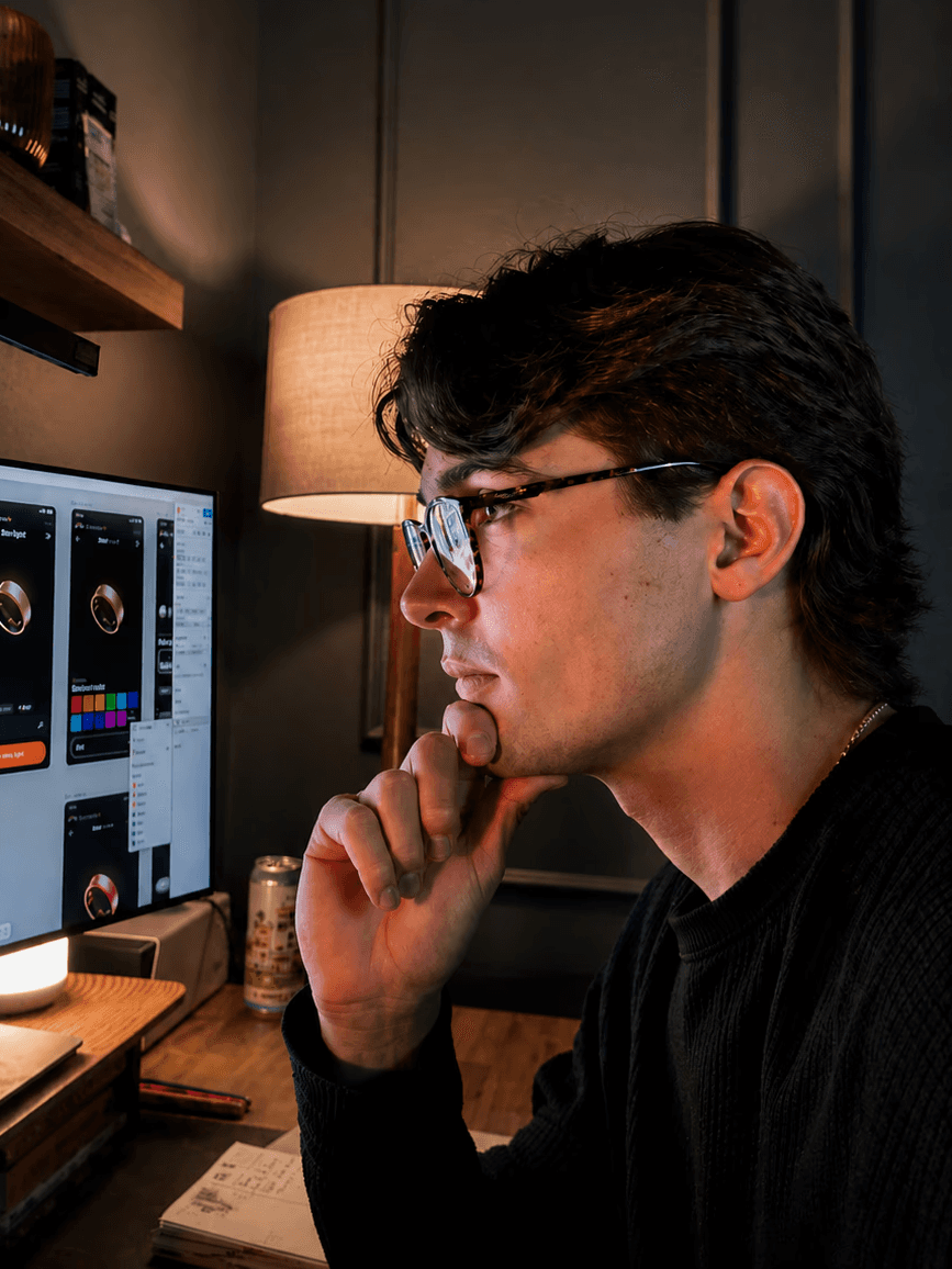 A young person with long hair and glasses sits at a desk looking at a monitor 