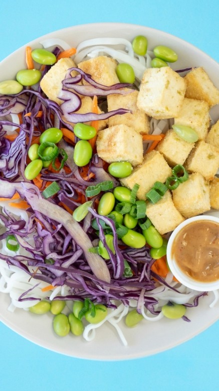 Bowl of Thai noodle salad with oven-roasted tofu and garnished with fresh vegetables and edamame beans on a blue background