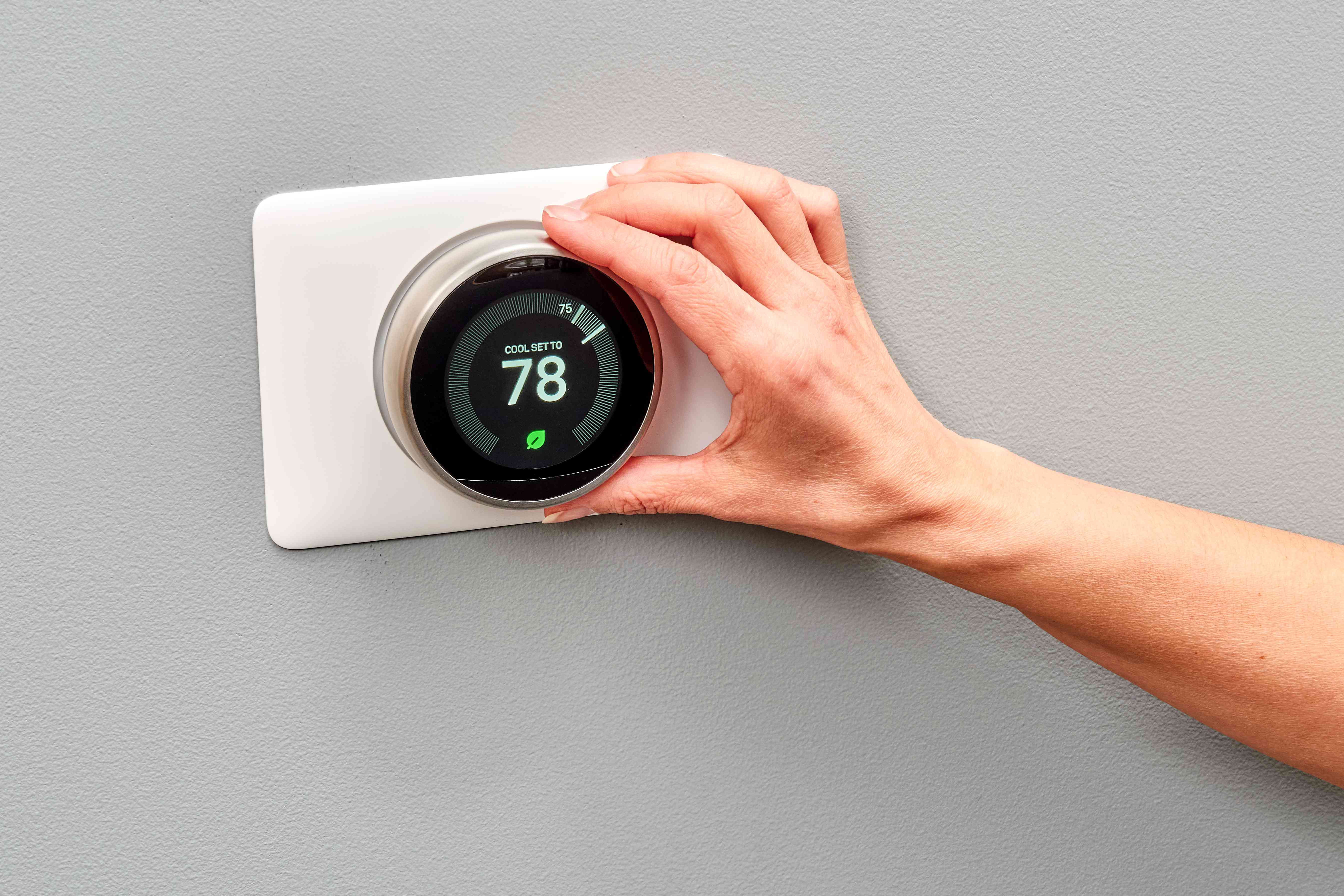 A hand adjusts a modern smart thermostat mounted on a wall. The black circular display shows the temperature set to 78 degrees Fahrenheit with a green leaf icon below the number.